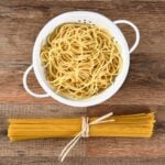 white colander of spaghetti with bundle of spaghetti on wooden board