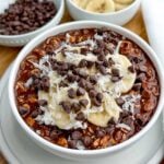 chocolate overnight oats with banana, coconut, and chocolate chips in a white bowl.