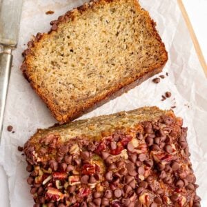 slice of chocolate chip pecan banana bread next to a cut loaf on white parchment papaer.