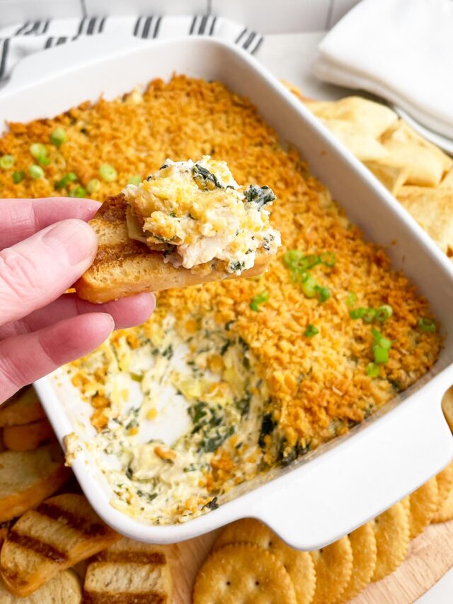 hand holding crostini with crab spinach artichoke dip on top with baking dish of dip in background
