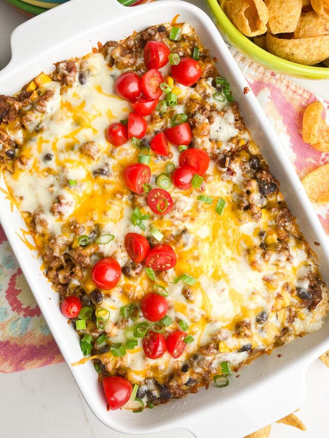 taco dip recipe with ground beef in white casserole dish with colorful napkin, plates, and scoops chips on counter