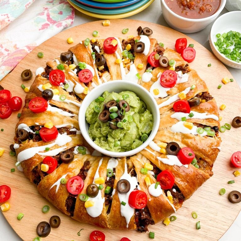 taco ring covered in toppings with guacamole in the center on a wooden cutting board
