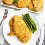 white plate with two baked thin chicken breasts and green beans with platter of chicken in background