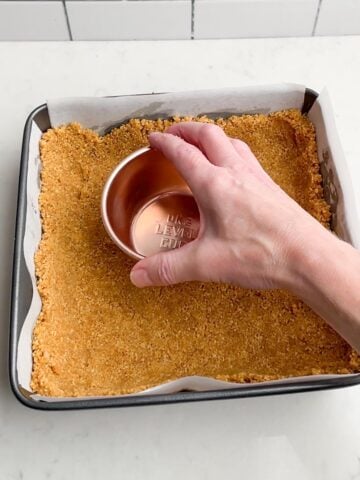 hand holding measuring cup pressing graham cracker crust into baking pan