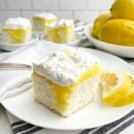 piece of lemon poke cake on white plate with bowl of lemons and plates of cake in background
