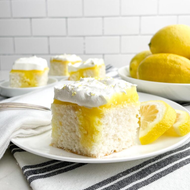 piece of lemon poke cake on white plate with bowl of lemons and plates of cake in background