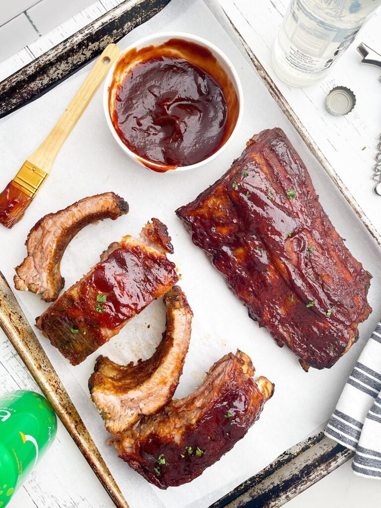 one full rack of ribs and individual ribs on white parchment with a bowl of BBQ sauce and a paint brush