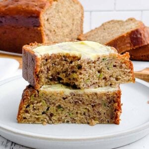 two half pieces of banana zucchini bread stacked on a plate with a loaf of banana zucchini bread in the background