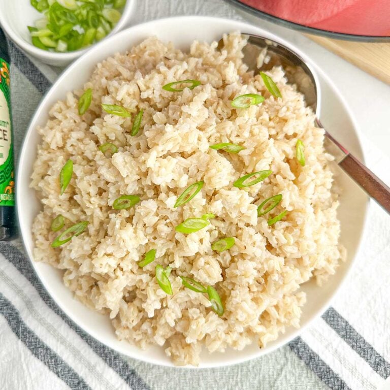 cooked brown rice and green onions in a white bowl by a red crock pot.