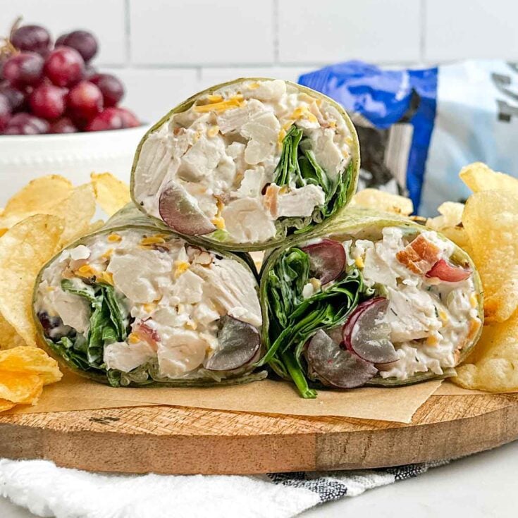 three chicken salad wrap half sandwiches on a wooden cutting board with kettle chips.