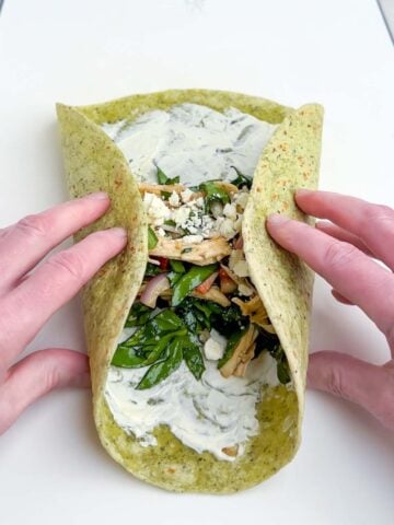 hands folding a chicken spinach wrap on a white cutting board.