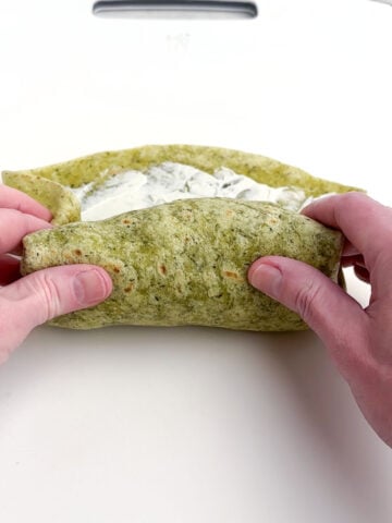 hands folding a chicken spinach wrap on a white cutting board.