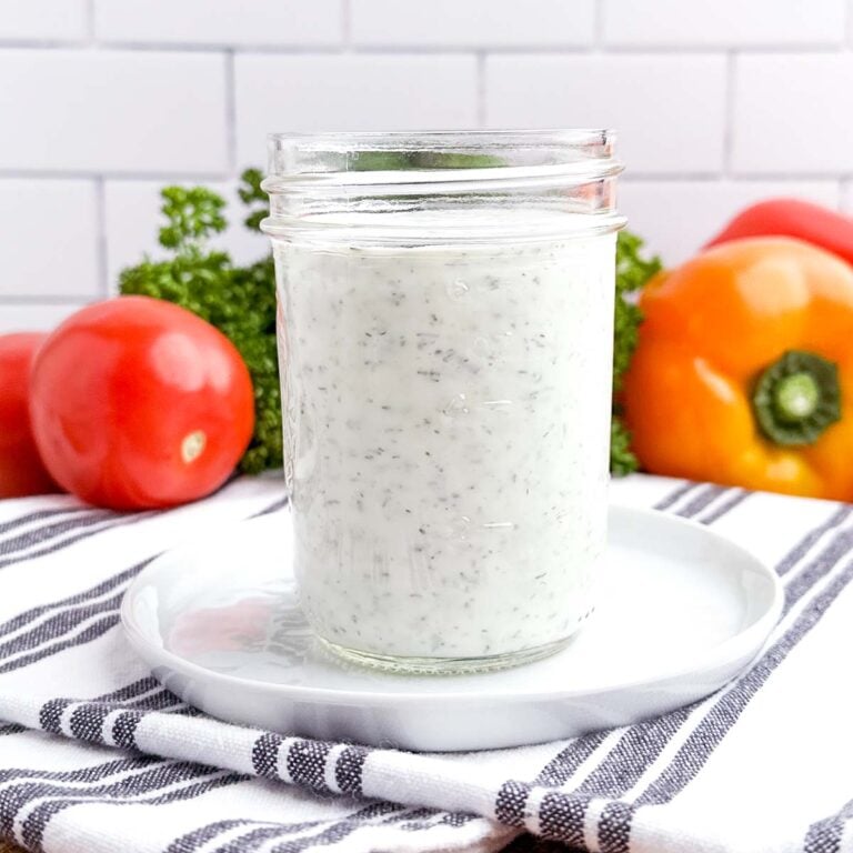 greek yogurt salad dressing in a clear jar on a striped tea towel surrounded by fresh vegetables.