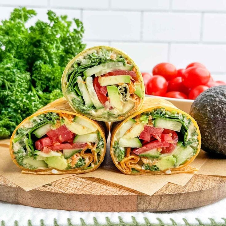 three raw veggie wrap halves stacked on a wooden cutting board.