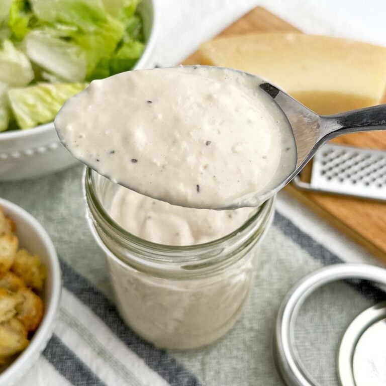 spoonful of caesar salad dressing in foreground with jar of dressing, lettuce, parmesan, and croutons in background.