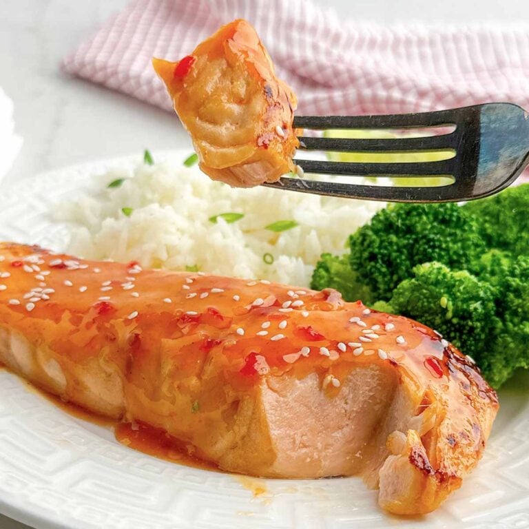 fork holding up a bite of sweet chili salmon in front of a white plate with rice and broccoli.