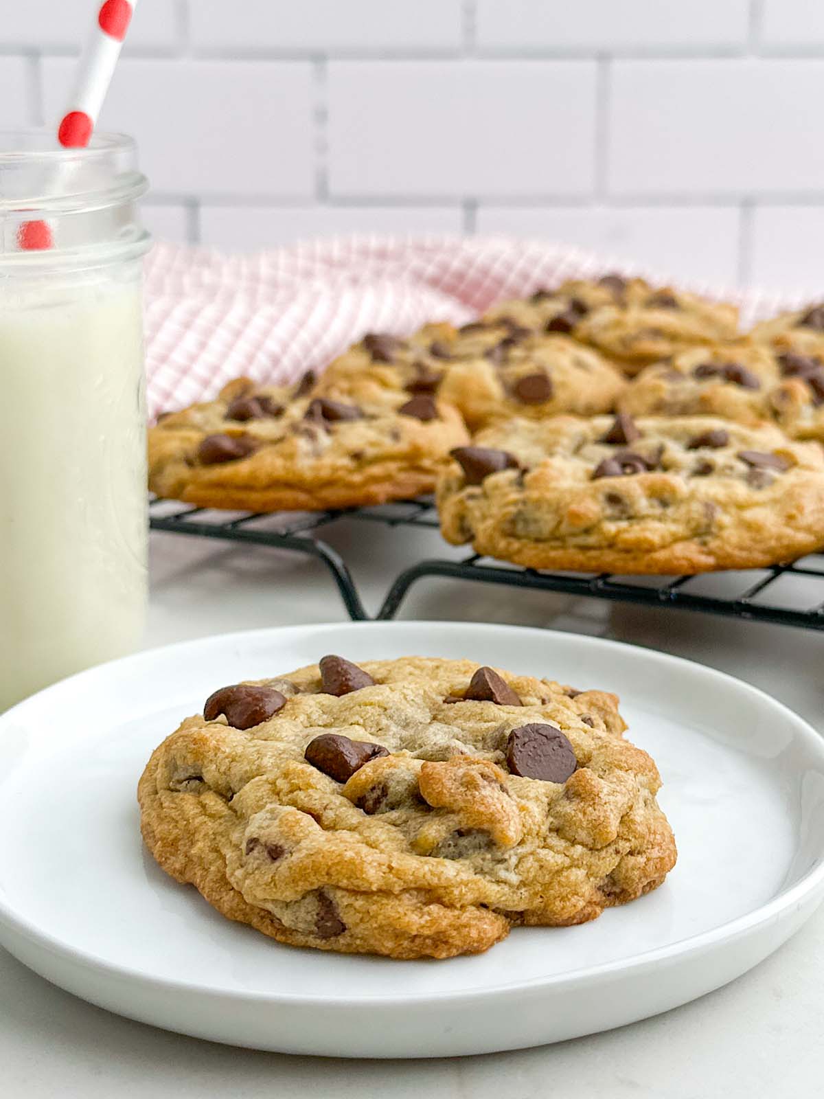chocolate chip cookie on a white plate next to a glass of milk and a wire rack of chocolate chip cookies.
