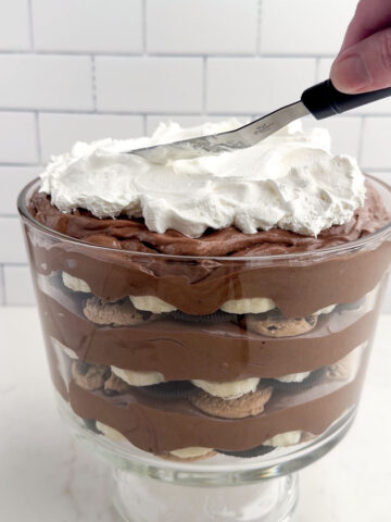 hand holding an offset spatula spreading cool whip on top of a layered banana chocolate pudding dessert in a clear trifle bowl.