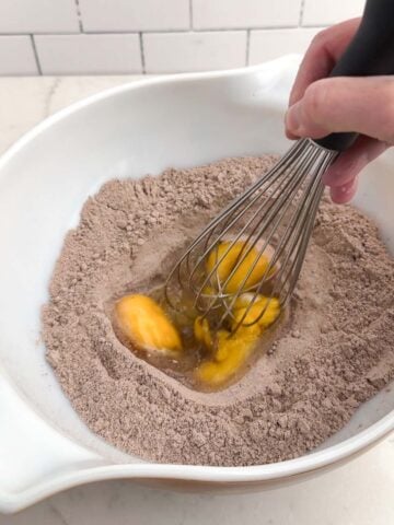 whisk beating eggs in the well of chocolate cake mix in a white bowl.
