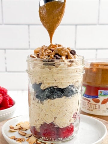 spoon drizzling melted almond butter over almond butter overnight oats with berries in a mason jar.