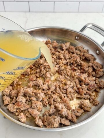 broth pouring from a measuring cup into a pan of cooked sausage.