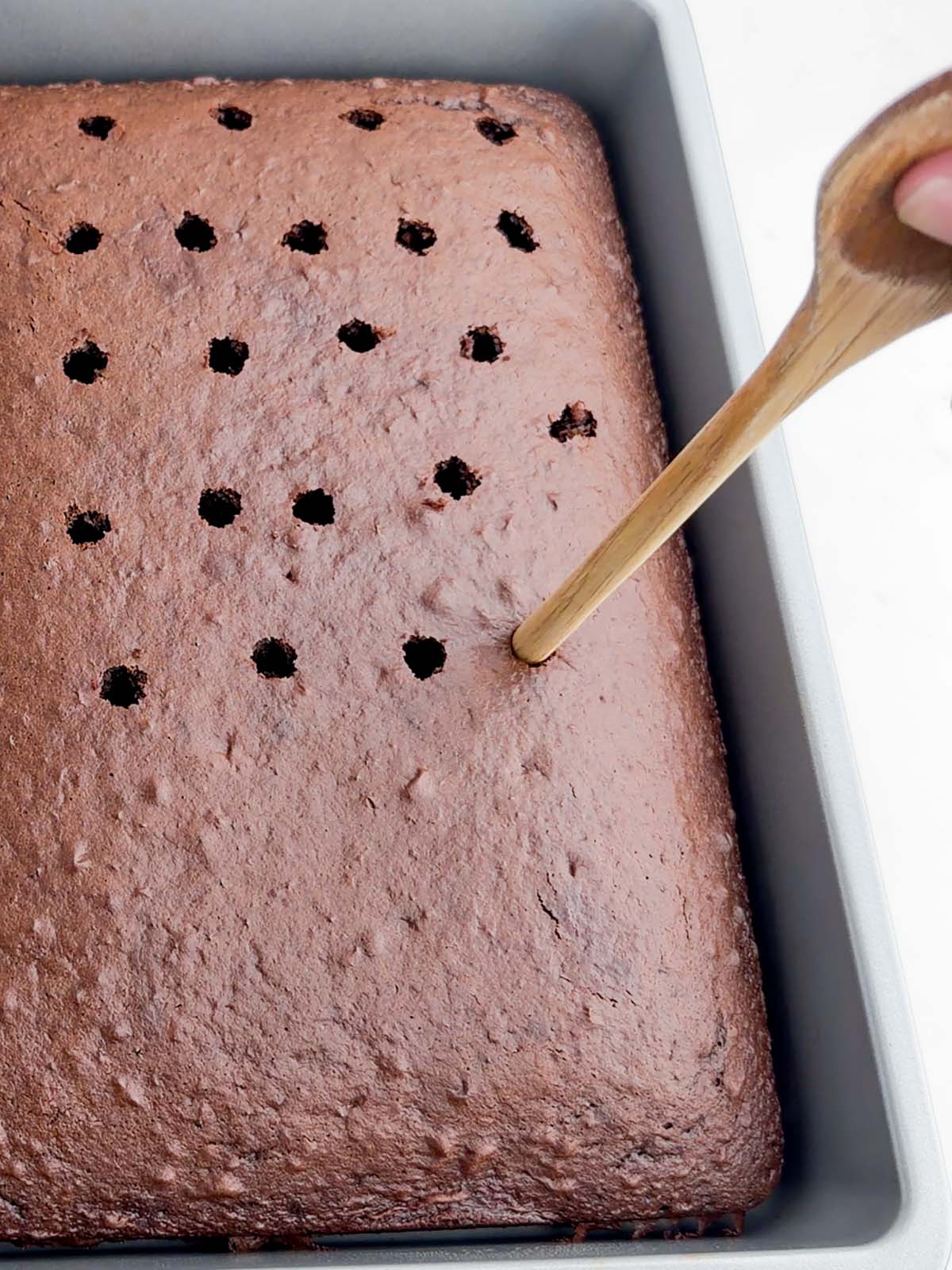poking holes in chocolate cake with the handle of a wooden spoon.